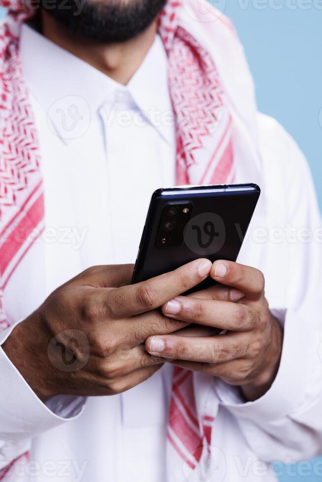 Arab man holding mobile phone in hands, scrolling social media network and texting online closeup. Muslim person in traditional clothes using smartphone and surfing internet photo