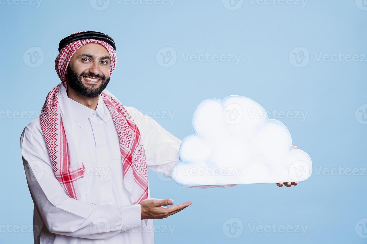 Carefree man wearing arabic traditional clothes posing with cloud shape cardboard banner studio portrait. Smiling muslim person holding showing white bubble and looking at camera photo