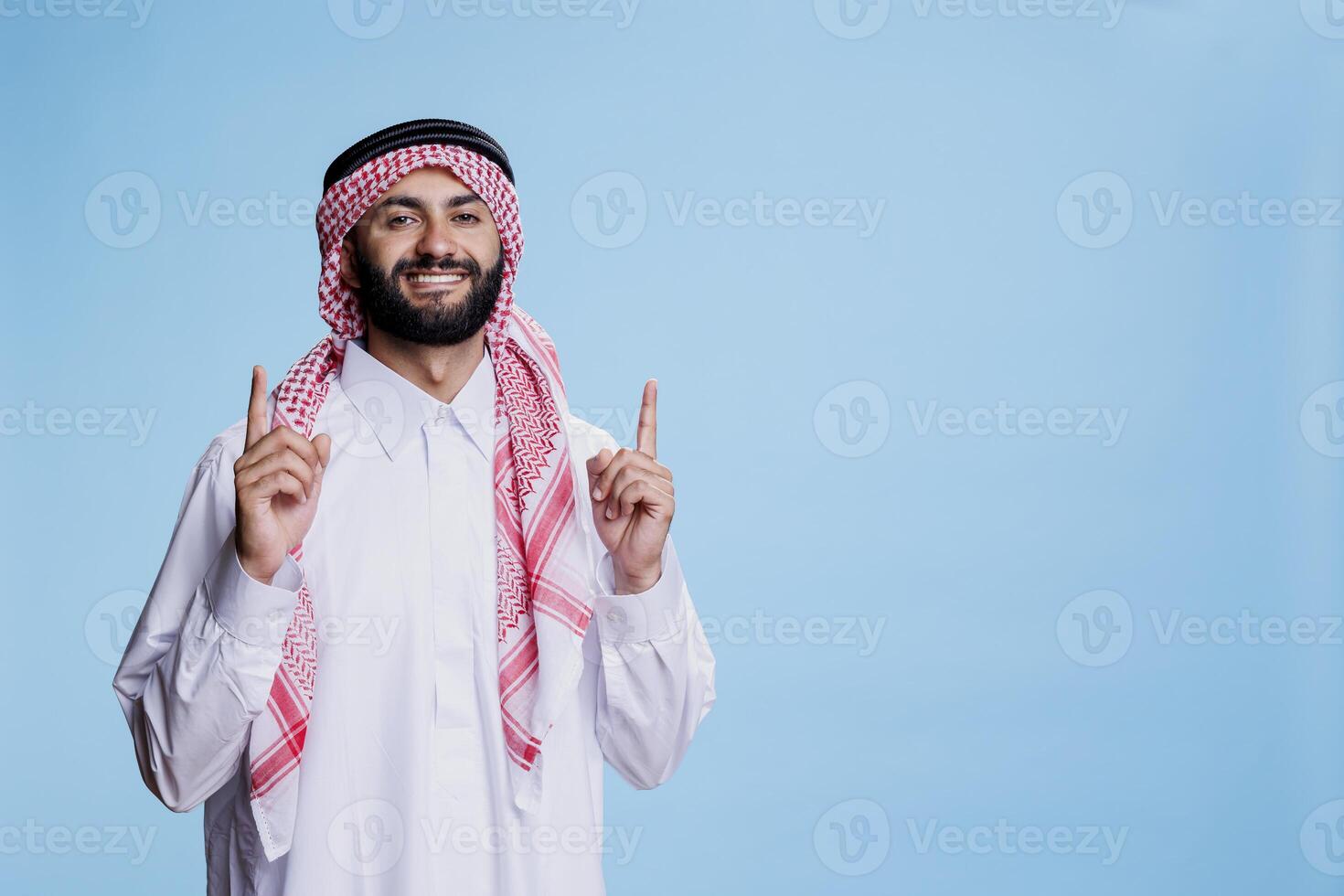 Excited muslim sales person pointing upwards direction while posing for studio portrait. Cheerful smiling arab man wearing traditional clothes showing up and looking at camera photo