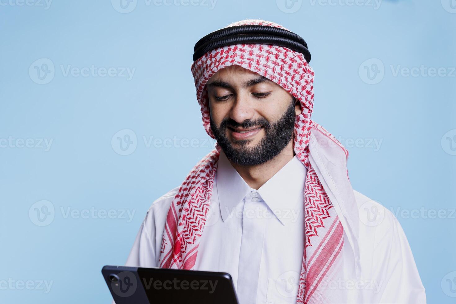 Smiling man wearing arabic clothes using digital tablet and scrolling social media. Cheerful muslim person dressed in ghutra headscarf and thobe tapping on portable device screen photo