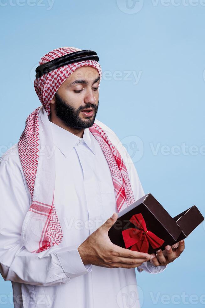 Muslim man wearing traditional attire holding giftbox with bow ribbon and opening festive present. Arab dressed in white thobe standing with giftbox in studio on blue background photo