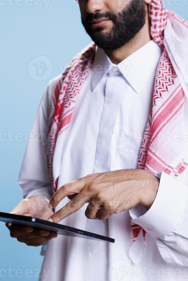 Man dressed in muslim white thobe and headscarf pressing on digital tablet touchscreen with fingers closeup. Arab wearing traditional clothes tapping on portable gadget screen photo