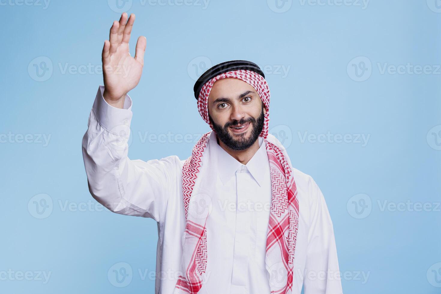 Smiling muslim man dressed in traditional clothes raising hand studio portrait. Islamic person greeting friend, waving with arm and looking at camera with cheerful expression photo