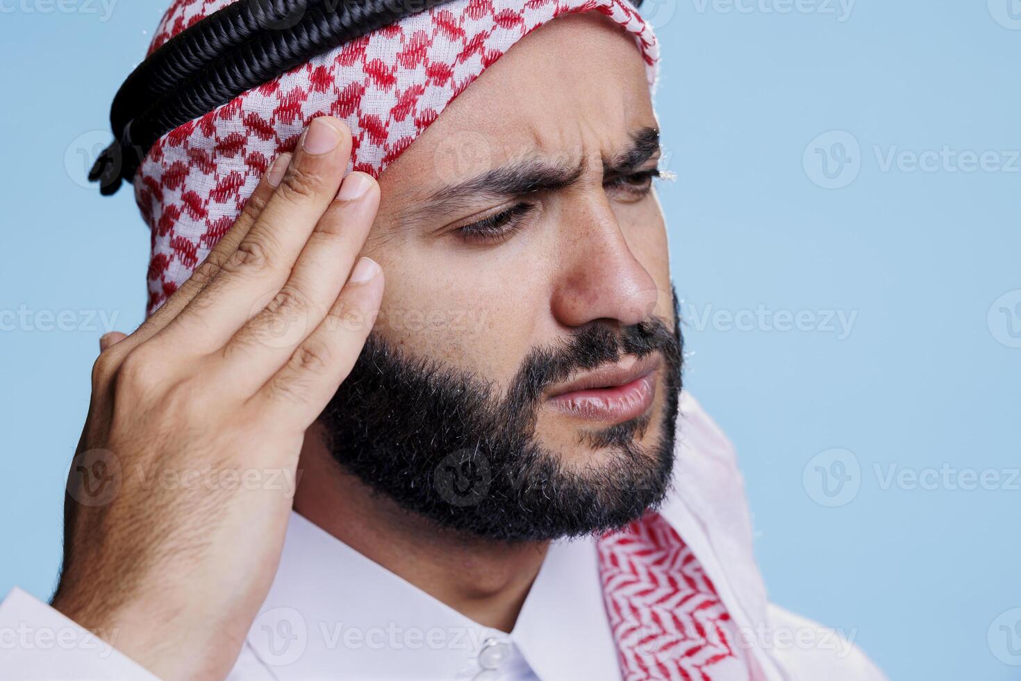 Muslim man rubbing temple and suffering from migraine symptom with tired expression. Arab person wearing thobe and ghutra traditional clothes having headache and feeling pain photo