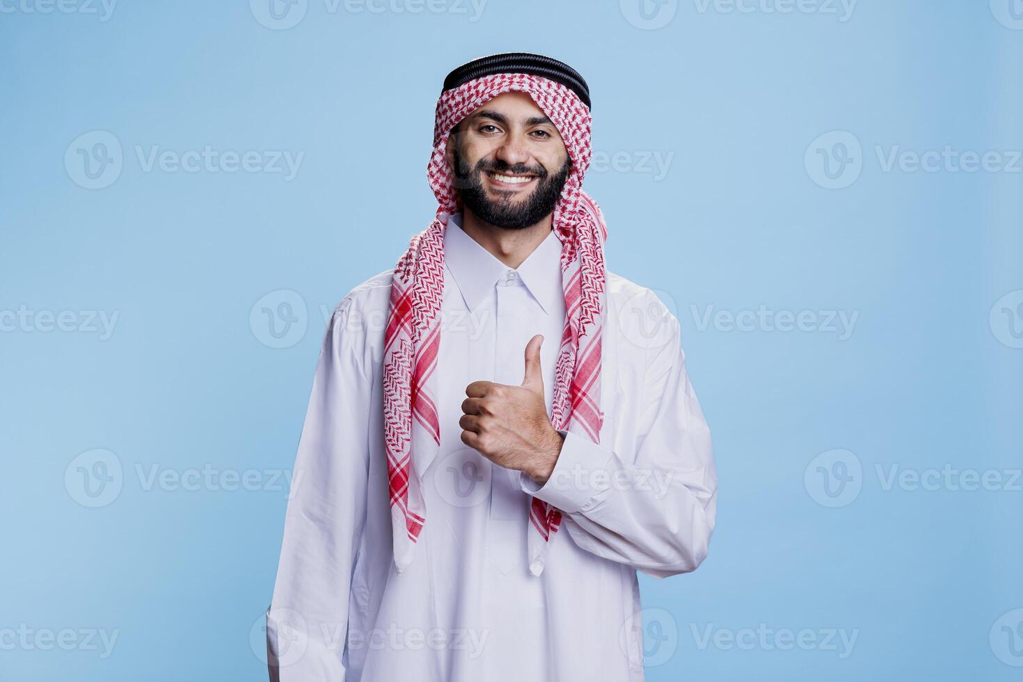 Arab man wearing islamic traditional clothes posing with thumb up signifying approval and agreement portrait. Smiling muslim person in thobe and ghutra showing like gesture photo