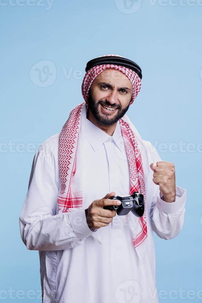 Excited muslim man holding joystick and showing clenched fist while winning game studio portrait. Happy person dressed in arabic traditional clothes posing with gamepad and winner gesture photo