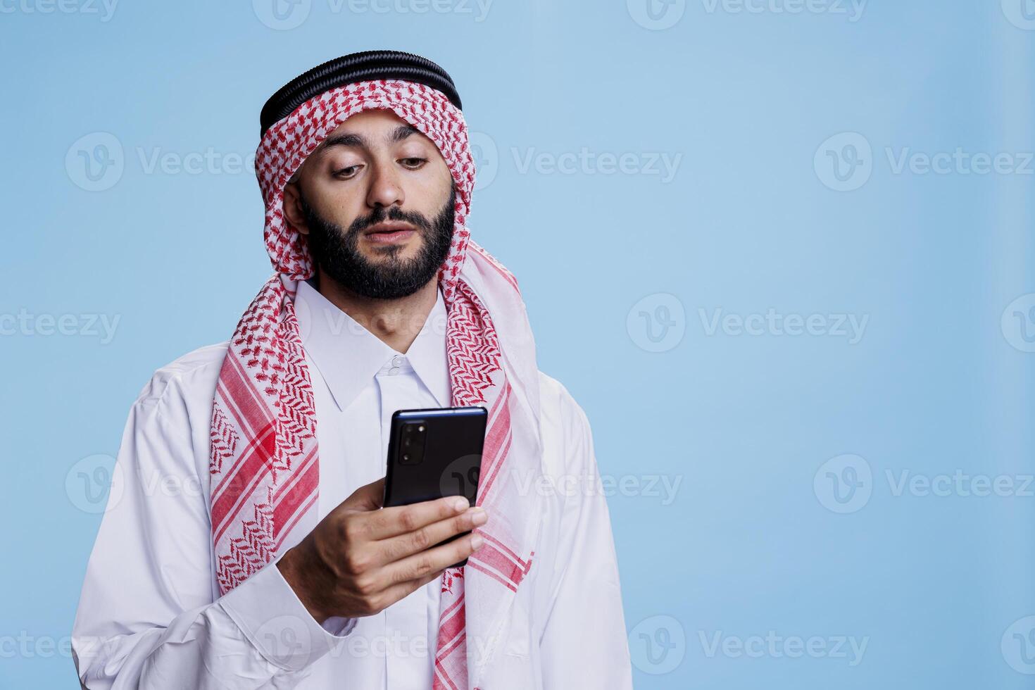 Muslim man wearing traditional clothes texting on smartphone while posing in studio. Arab person dressed in thobe and ghutra sending online message on mobile phone on blue background photo