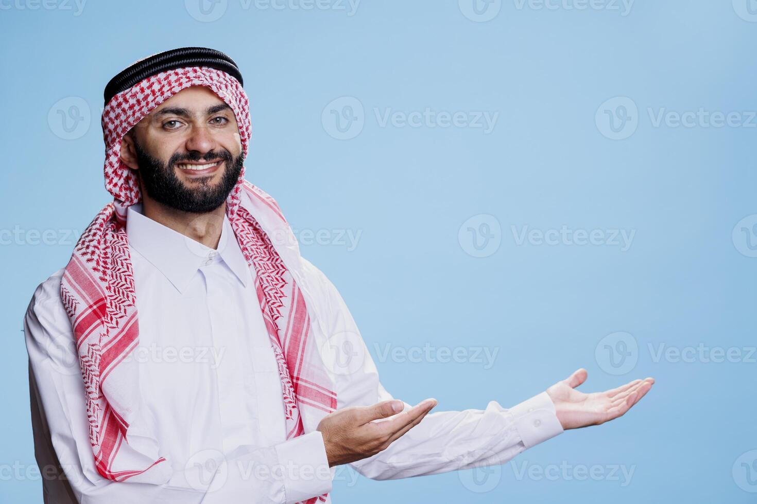 Smiling muslim man showcasing product by pointing to side with arms and looking at camera with cheerful expression. Arab dressed in islamic thobe and headscarf, pointing at left direction photo