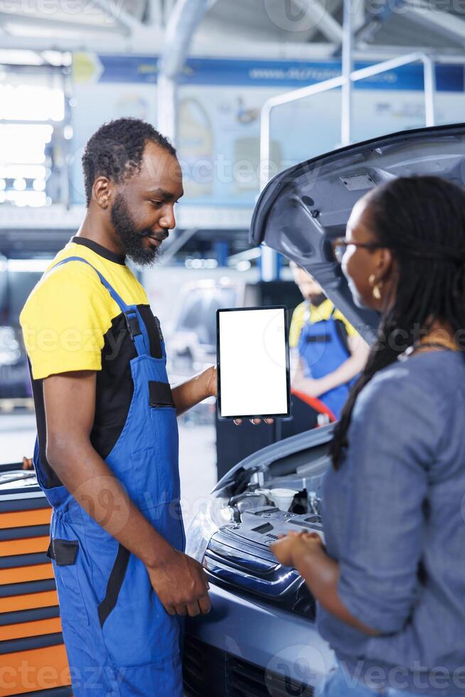 Smiling engineer in car service uses mockup tablet to order new compressor belt for damaged vehicle. Employee showing client needed components replacement on isolated screen device photo