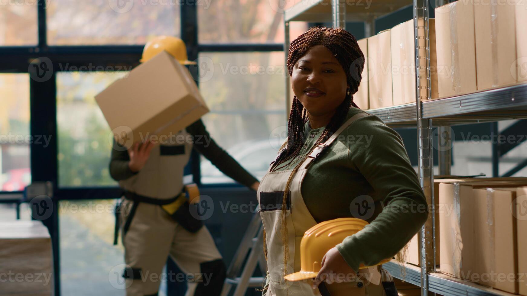 Portrait of cheerful african american warehouse sorter in protective uniform dealing with packaged stock in cardboard boxes ready to be shipped, happy to work in professional distribution center photo