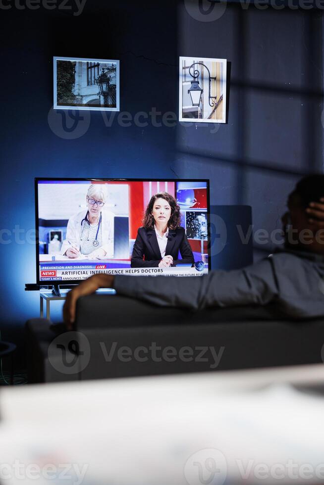 Curious spectator reads news coverage titles on television, sits on couch and listens to journalist lay out recent global events. Young person watching daily news headlines, tv content. photo