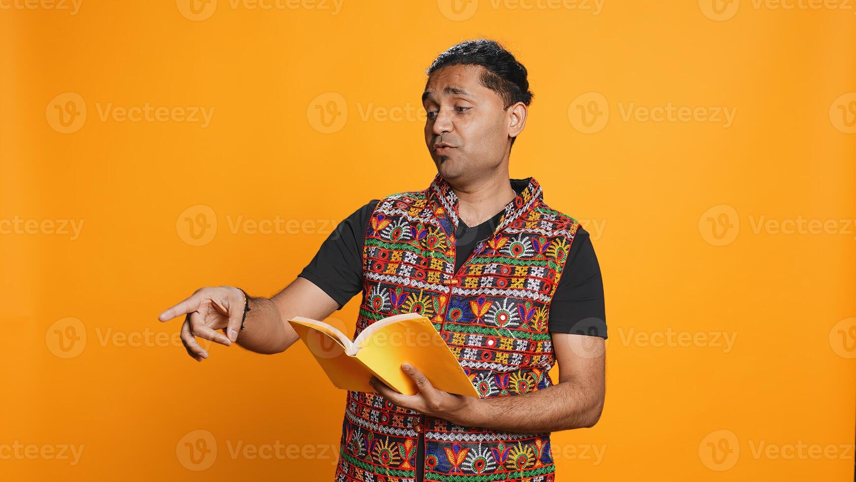 Joyous indian teacher reading book for pupils during class, isolated over studio background. Cheerful man holding novel, telling story to kindergarten kids, teaching them, camera B photo