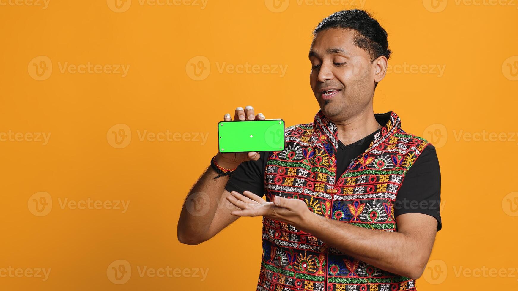 Content creator presenting green screen mobile phone, isolated over studio background. Indian person reviewing copy space mockup smartphone device for internet channel, camera A photo