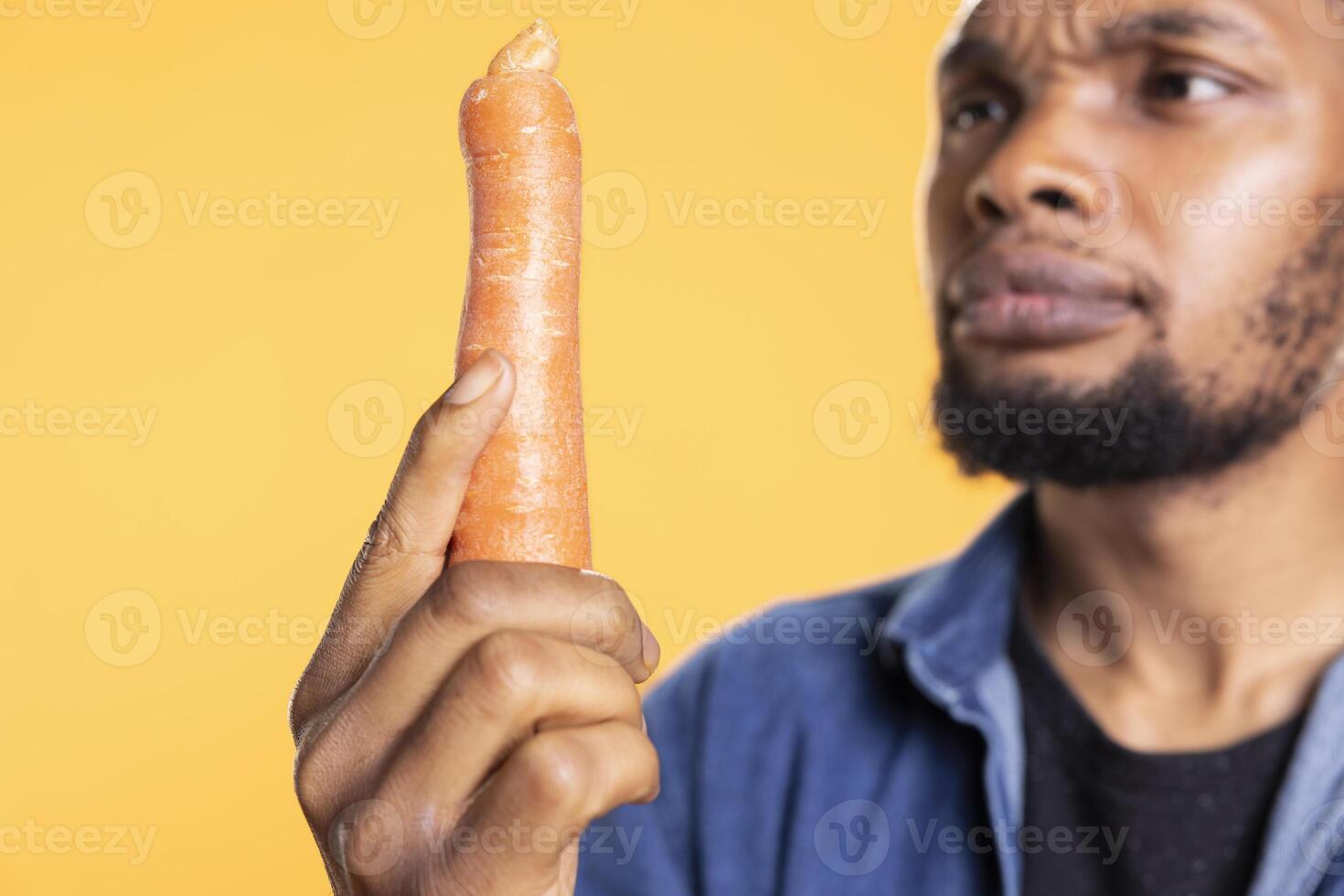 African american guy in doubt inspecting an organic carrot, looking intensely at additives free freshly harvested vegetable. Young person examining natural locally grown veggie, zero waste. photo