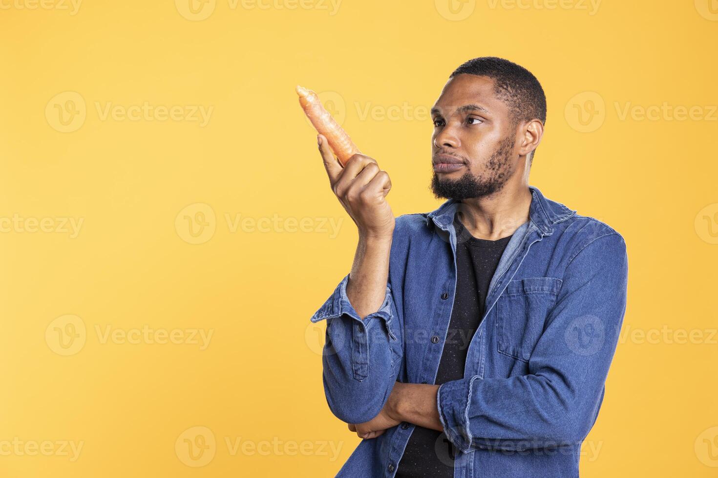 Happy charismatic person admiring ethically sourced carrots in studio, enjoying vegan nutrition and healthy eating. African american joyful guy smiling at organic ripe vegetables. photo