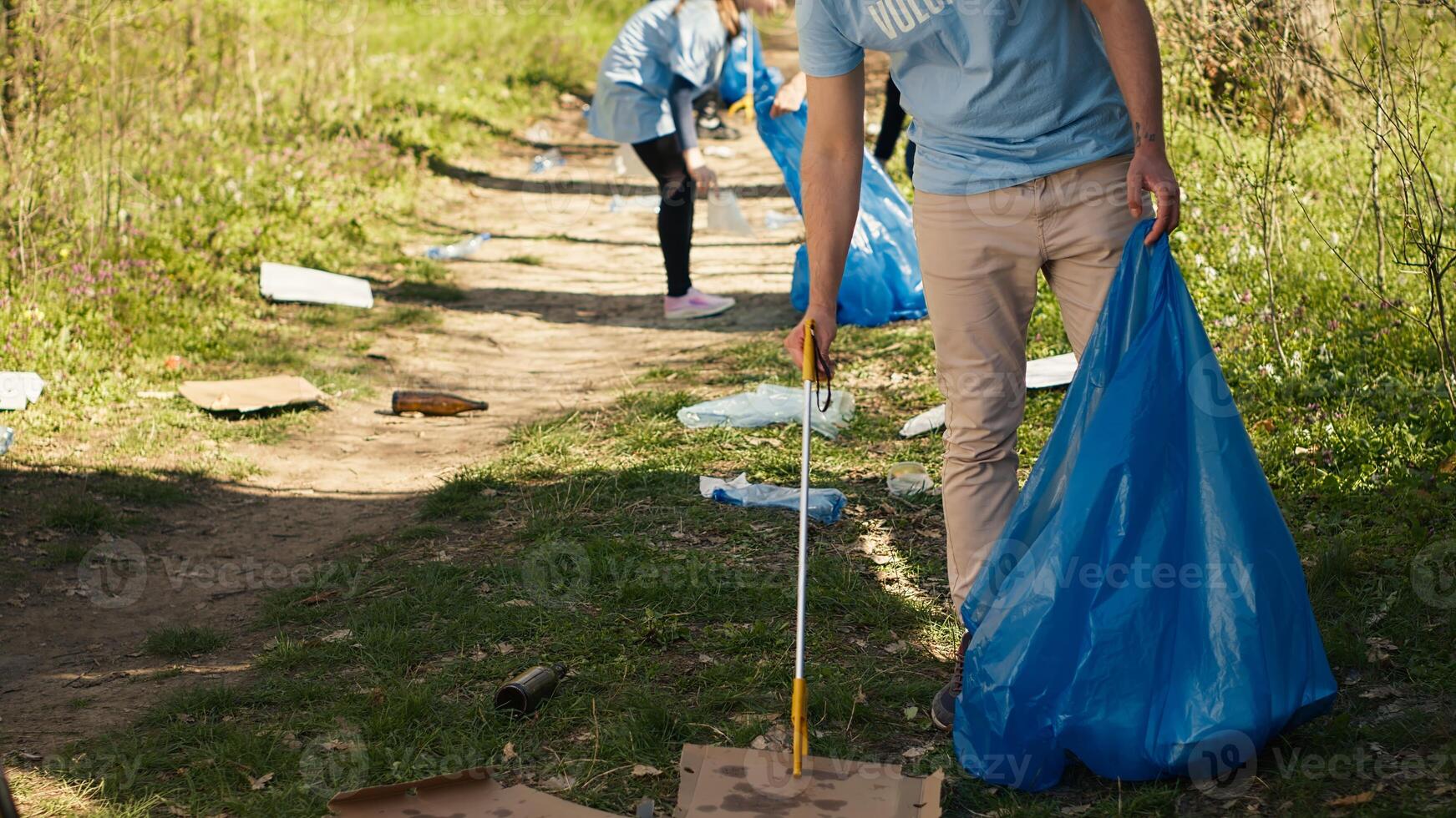 Young volunteer doing litter cleanup with a long claw to grab trash and store it in a garbage bag, dealing with pollution an illegal dumping problem. Activist protecting natural ecosystem. Camera A. photo