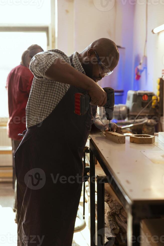 African american carpenter drills hole into timber block, doing furniture assembling in studio. BIPOC woodworking specialist inserting nail into piece of wood using electric power drill tool photo