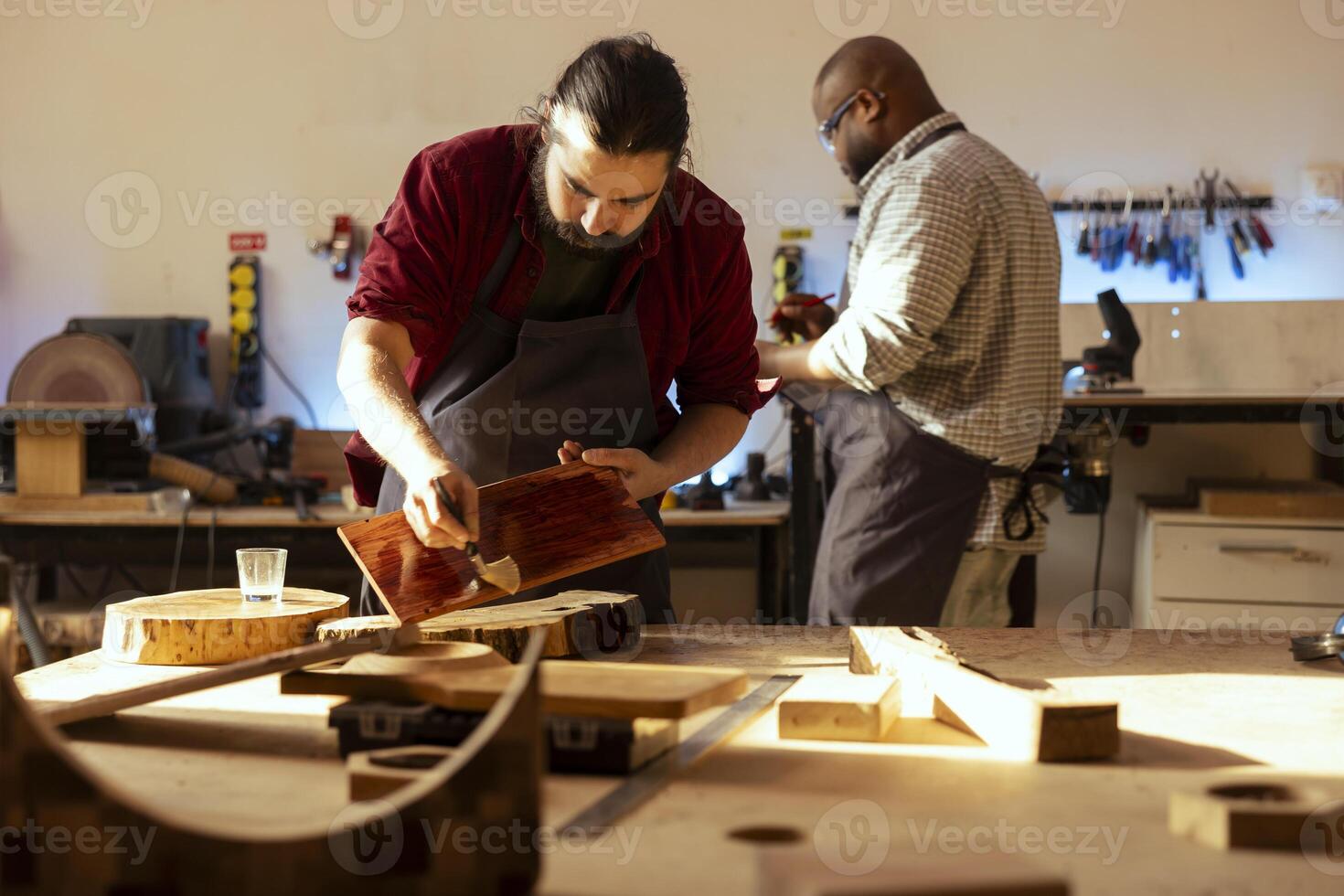 Carpenter brushing varnish on wood to build up protective layer with coworker in background solving tasks. Woodworker lacquering plank after sanding surface next to colleague working photo