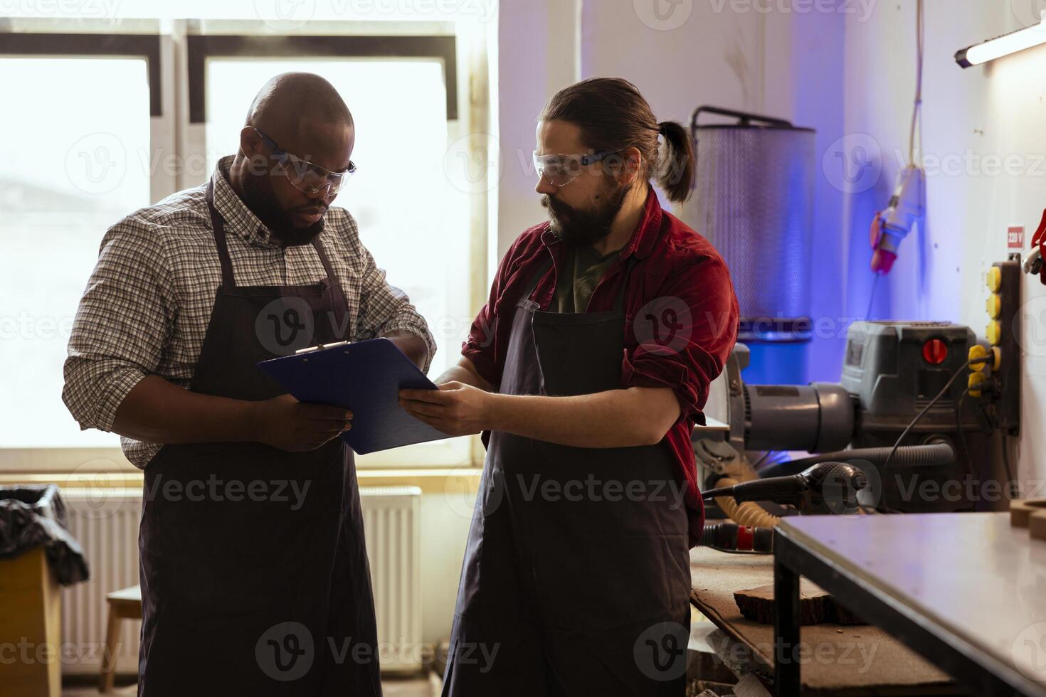 Teamworking carpenters wearing safety goggles drawing blueprints on notepad to make creative wood art pieces. Artisan coworkers looking at woodworking projects, using protective equipment photo