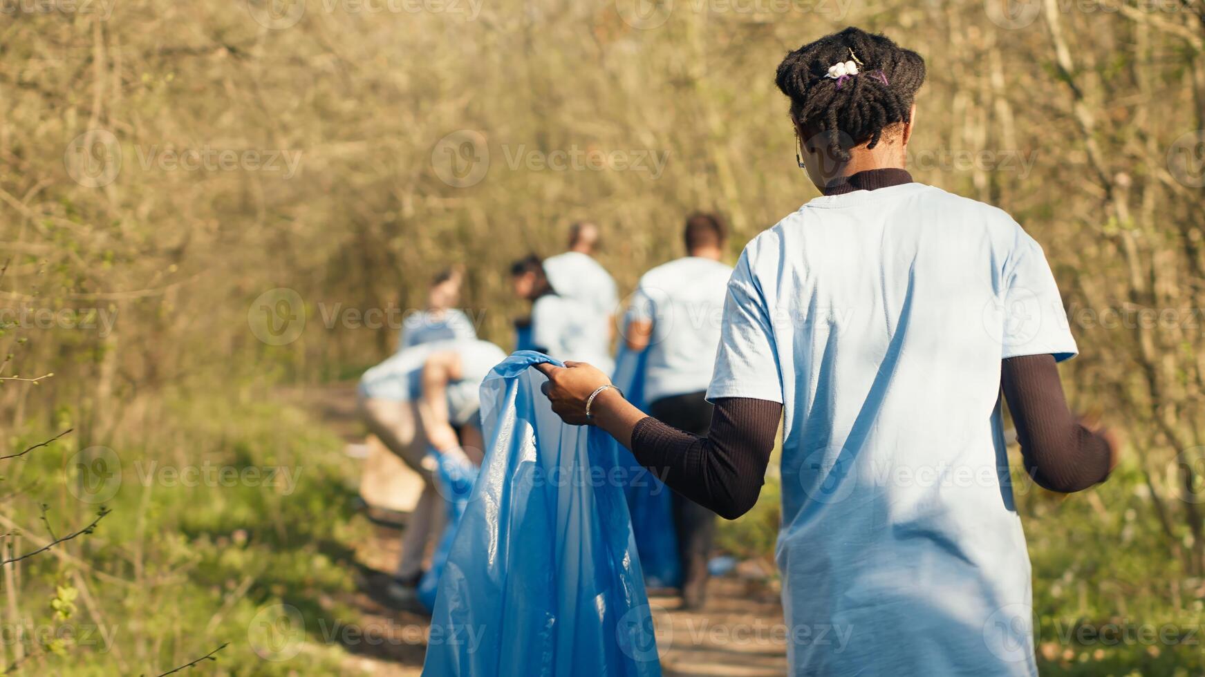African american girl collecting rubbish in a trash bag using tongs, recycling plastic waste and picking up garbage in a forest. Young woman activist clearing the woods from junk. Camera A. photo