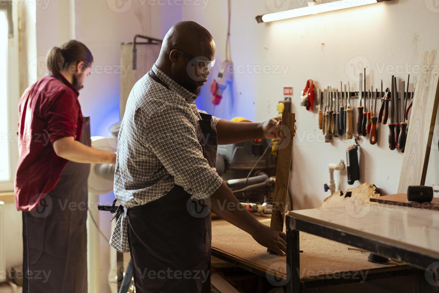 African american manufacturer wearing safety goggles while working with wood materials to avoid injury. Cabinetmaker using protective equipment to prevent workplace injuries while holding lumber piece photo