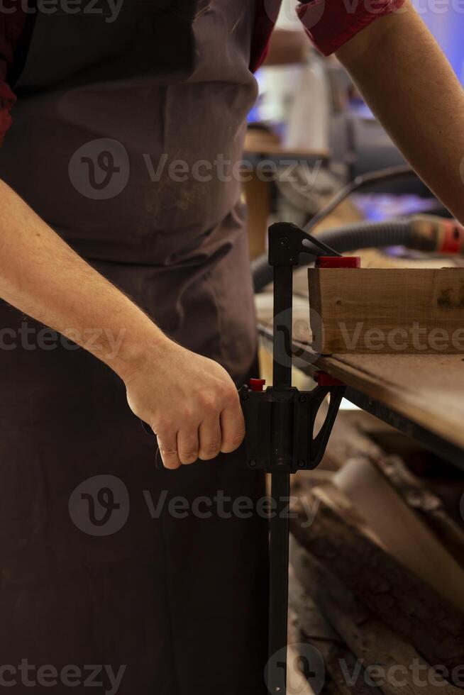 Artisan using bench vise to hold lumber block, starting designing wood art in studio, close up shot. Woodworking specialist in carpentry shop using sash clamp to secure piece of wood tightly photo