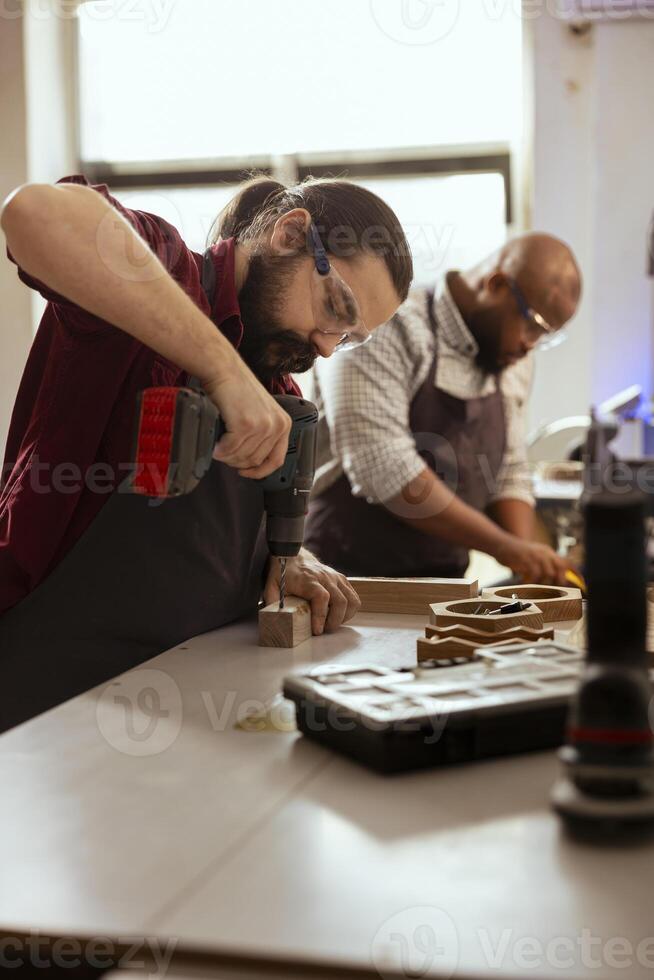 Woodworker next to BIPOC coworker using power drill to create holes for dowels in wooden board. Carpenter working with apprentice, sinking screws into wooden surfaces with electric tool photo