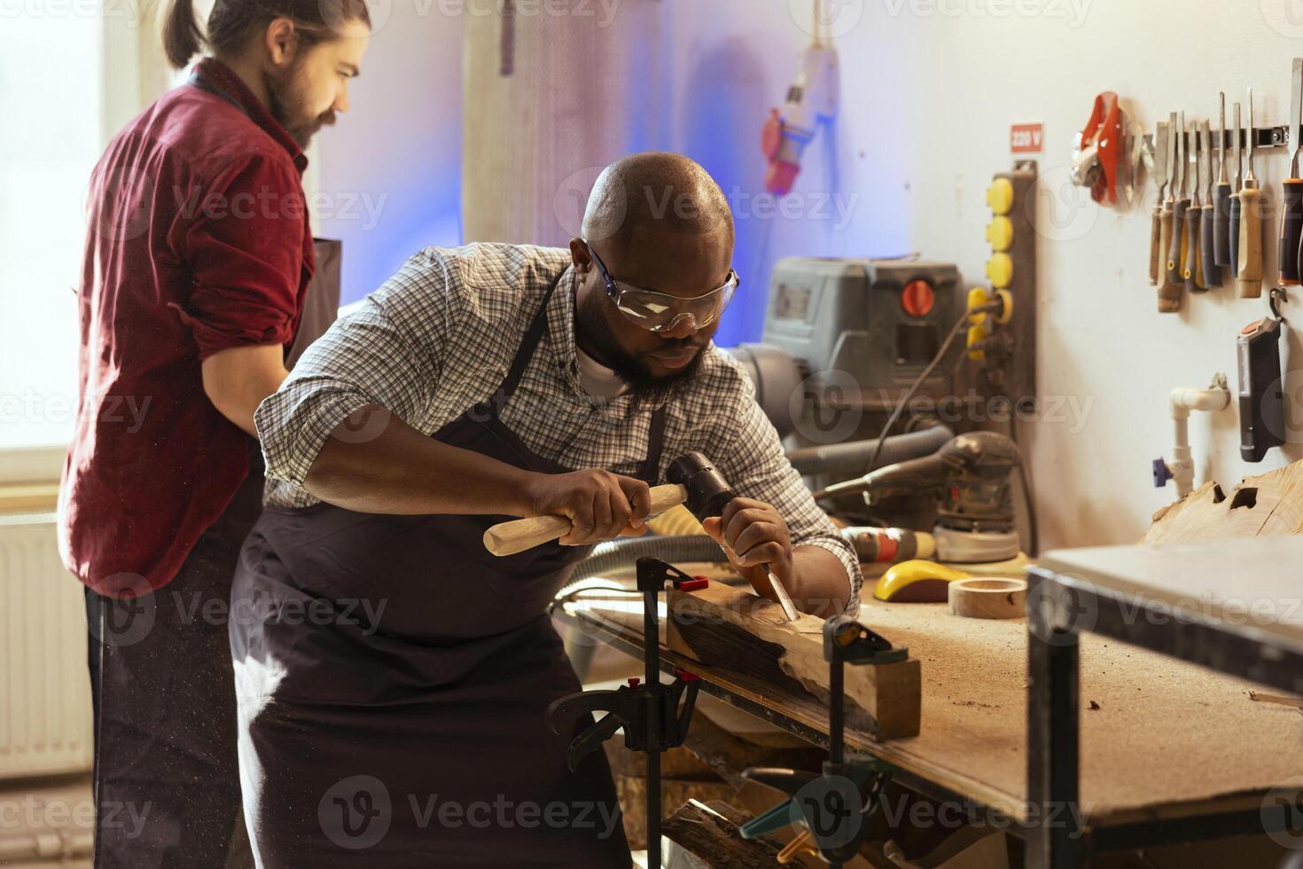 African american man wearing protection equipment while carving wood to prevent workplace accidents. Cabinetmaker equipped with safety glasses while using chisel and hammer, sculpting wooden surface photo