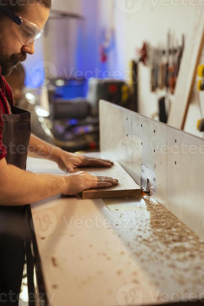 Carpenter puts wood block through spindle moulder wearing safety glasses to avoid injury. Cabinetmaker using heavy machinery to craft furniture using protective gear to prevent accidents photo