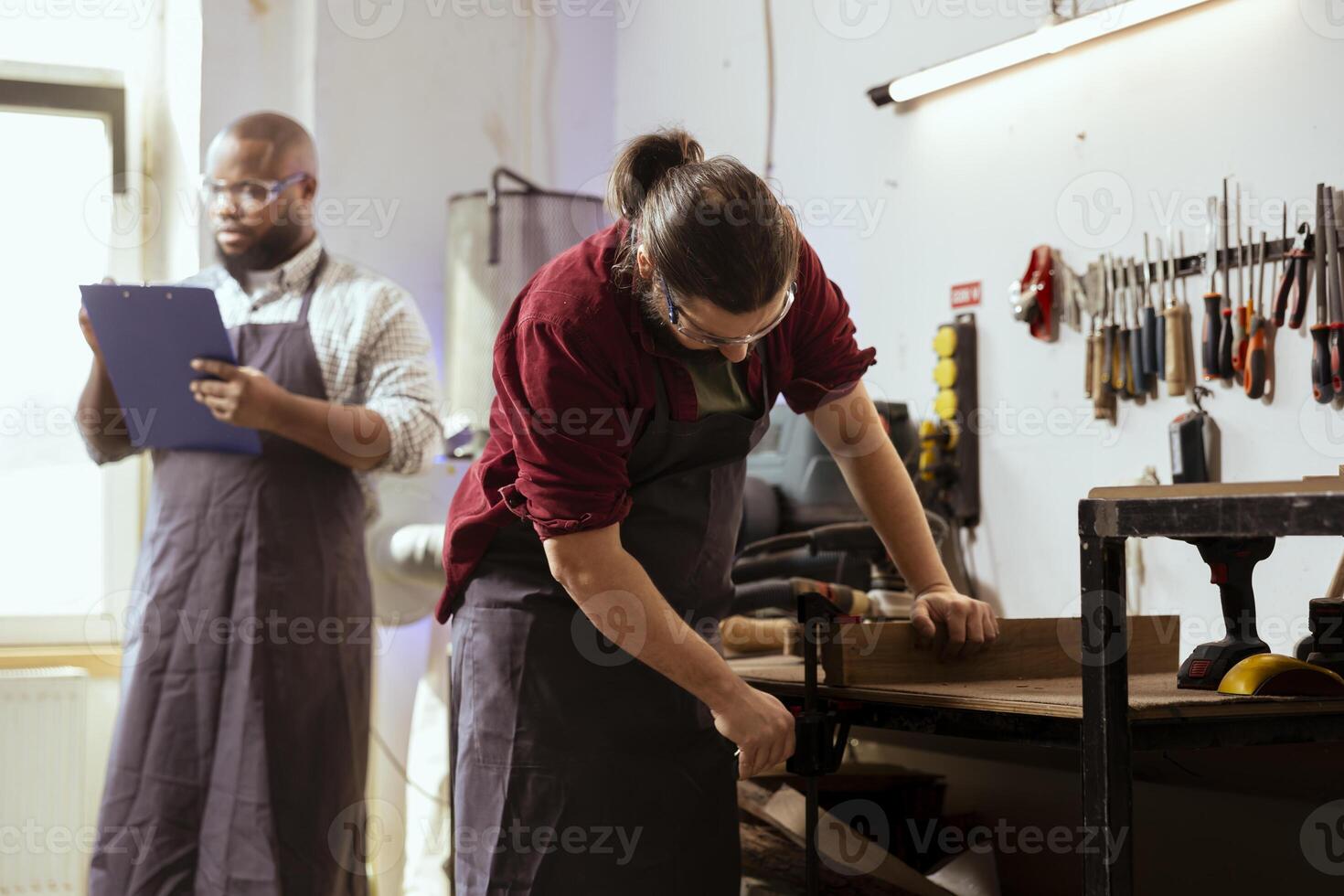 Woodworker using bench vise to hold lumber block, starting furniture assembling in workshop next to BIPOC coworker. Craftsperson using vice tool to clamp piece of wood with african american colleague photo