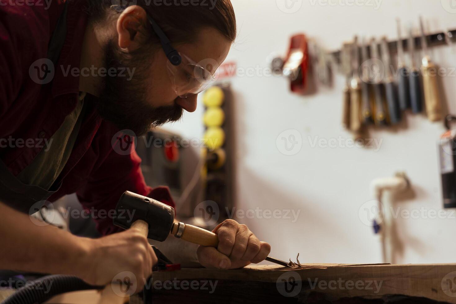 Sculptor shaping raw timber using chisel and hammer in carpentry shop, creating wood art, wearing safety glasses. Artist making wood sculptures, engraving lumber with tools, using protective gear photo