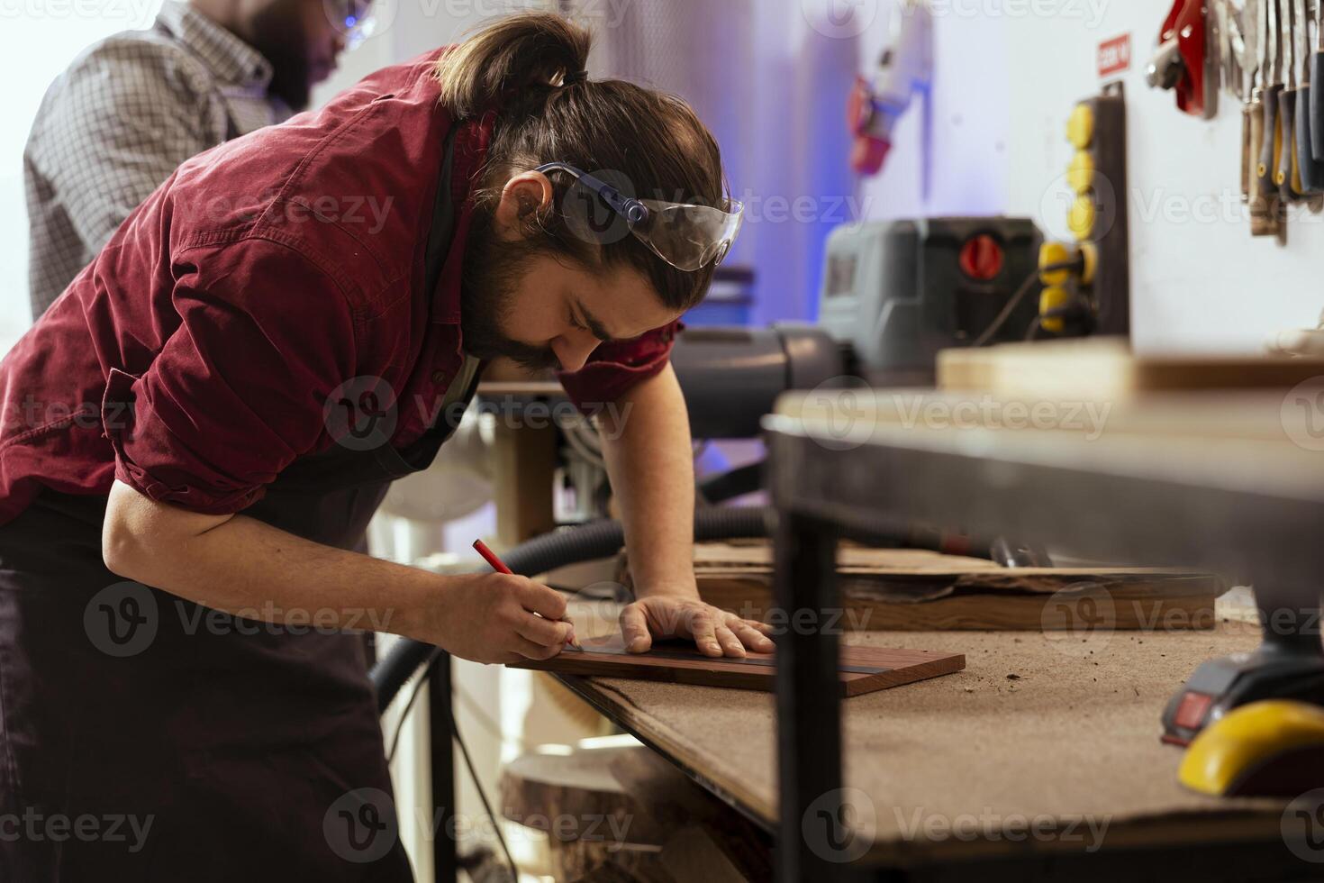 Craftsperson using ruler to take measurements of wood plank with coworker in background solving tasks. Man drawing with pencil on wooden board to determine where to make cut near BIPOC colleague photo