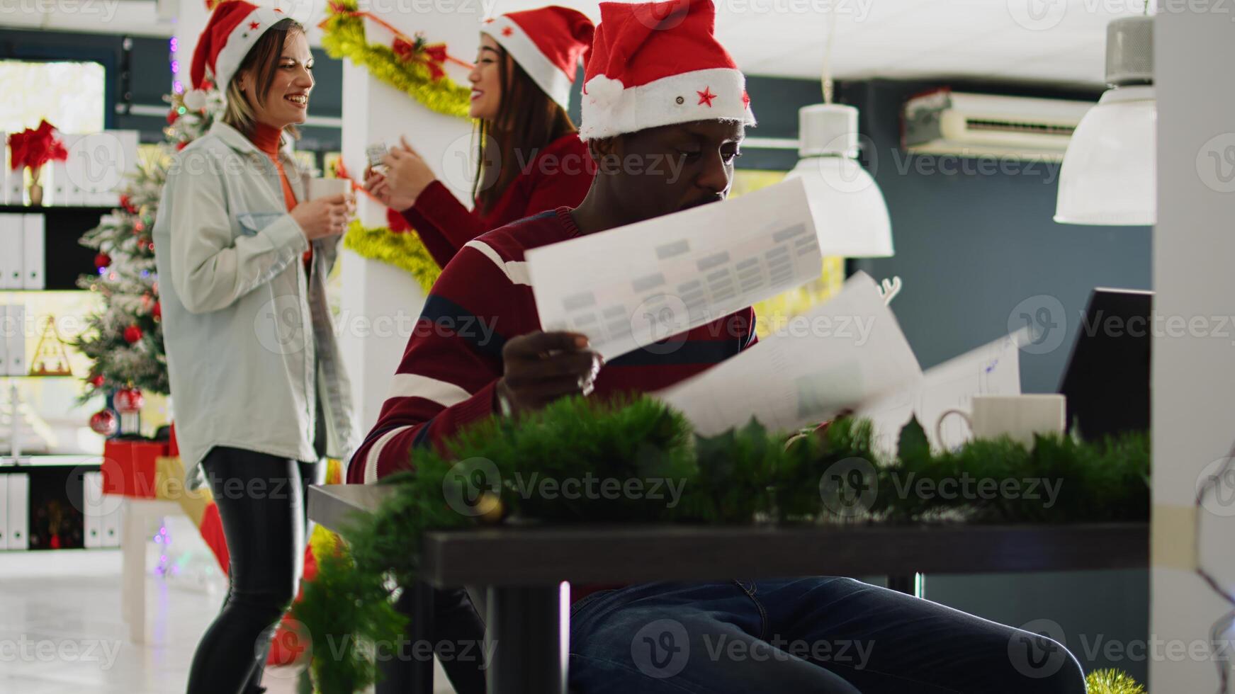African american employee in Christmas decorated office working on overdue project while carefree coworkers enjoy coffee break together. Worker frustrated by women laughing and distracting him photo