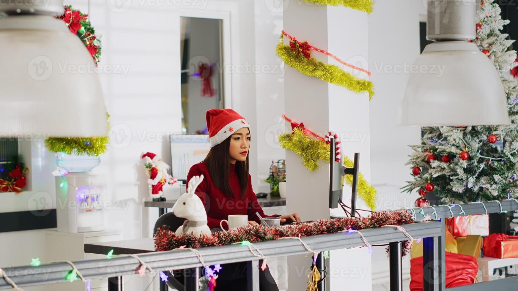 Proficient asian employee wearing Santa hat working in Christmas ornate office. Adept worker looking over business revenue numbers at computer desk in diverse xmas ornate workplace photo