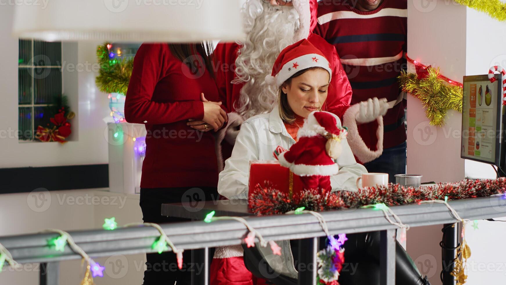 Manager dressed as Santa during Christmas season delegates project tasks to workers. Supervisor in xmas decorated office discussing with employees during winter holiday season, handheld camera photo