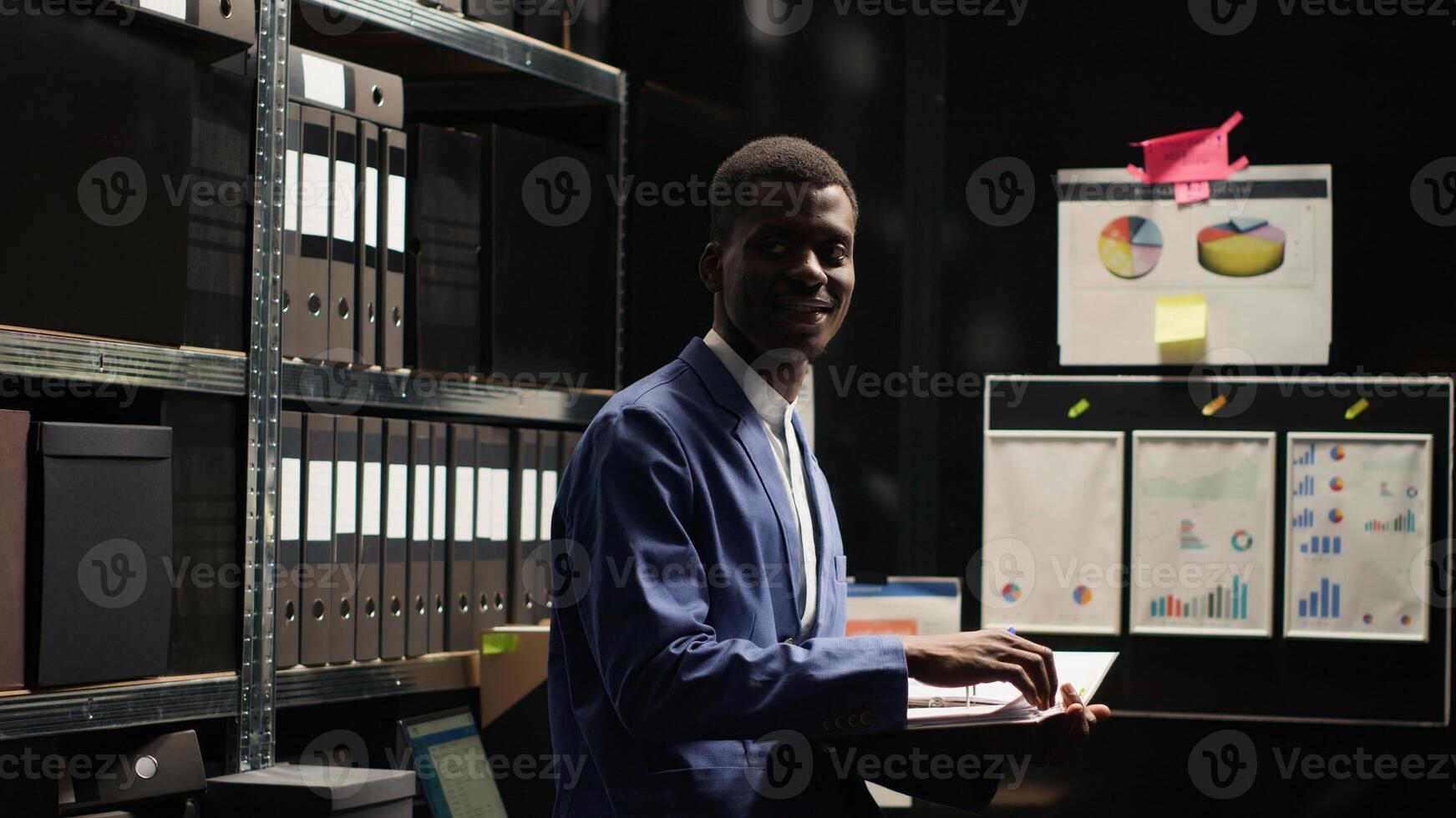 Portrait shot of african american investigator standing and reviewing case files in evidence room. Black man in dimly lit office checking suspect statements and records. Slow-mo, looking at camera. photo