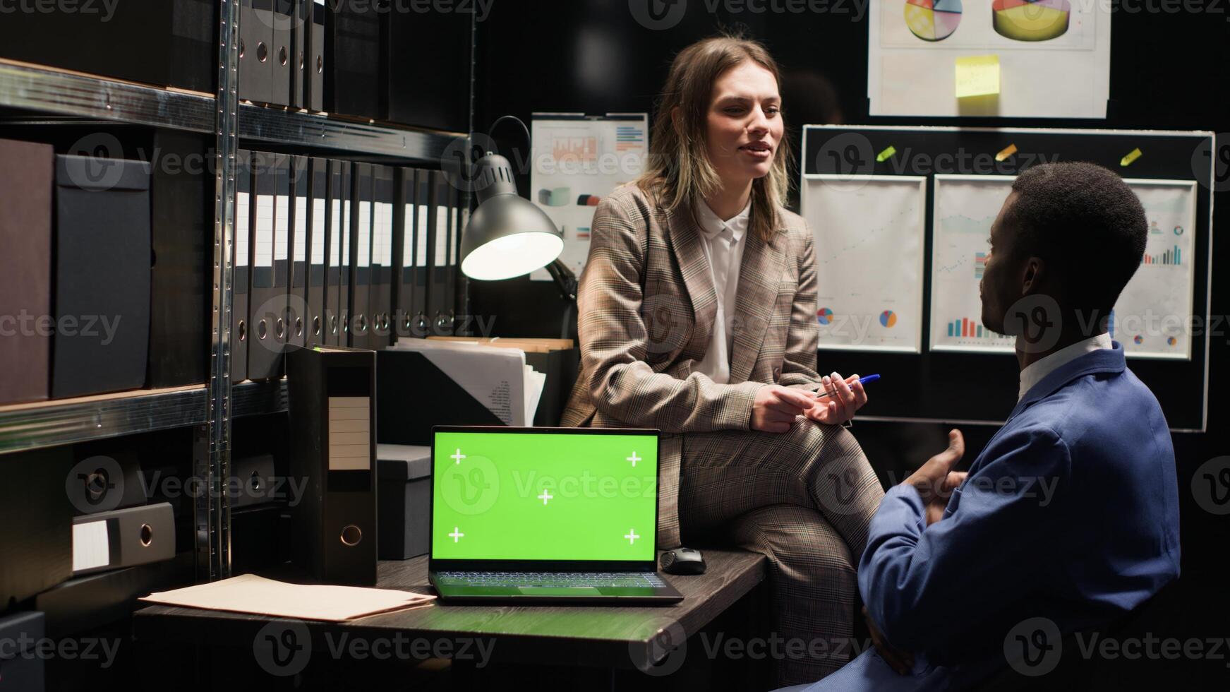 Policewoman talks with black man drinking coffee in office, with laptop displaying greenscreen placed on table. Wireless computer with isolated mockup template on desk while detectives discuss a case. photo
