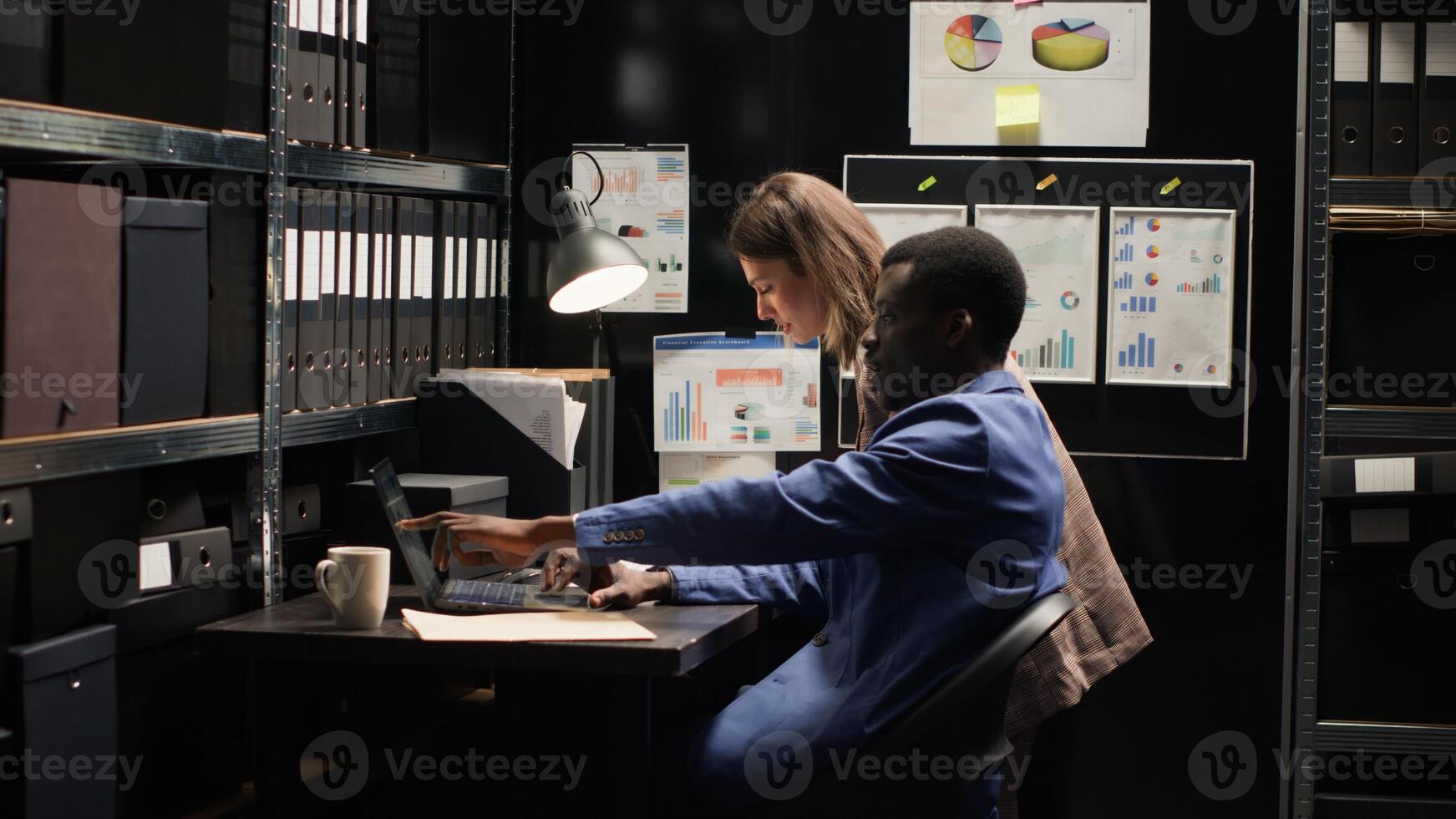 Diverse detectives in well-organized office reviewing evidence, surrounded by shelves filled with files. Two law enforcement officers with laptop, diligently investigate criminal profiles and records. photo