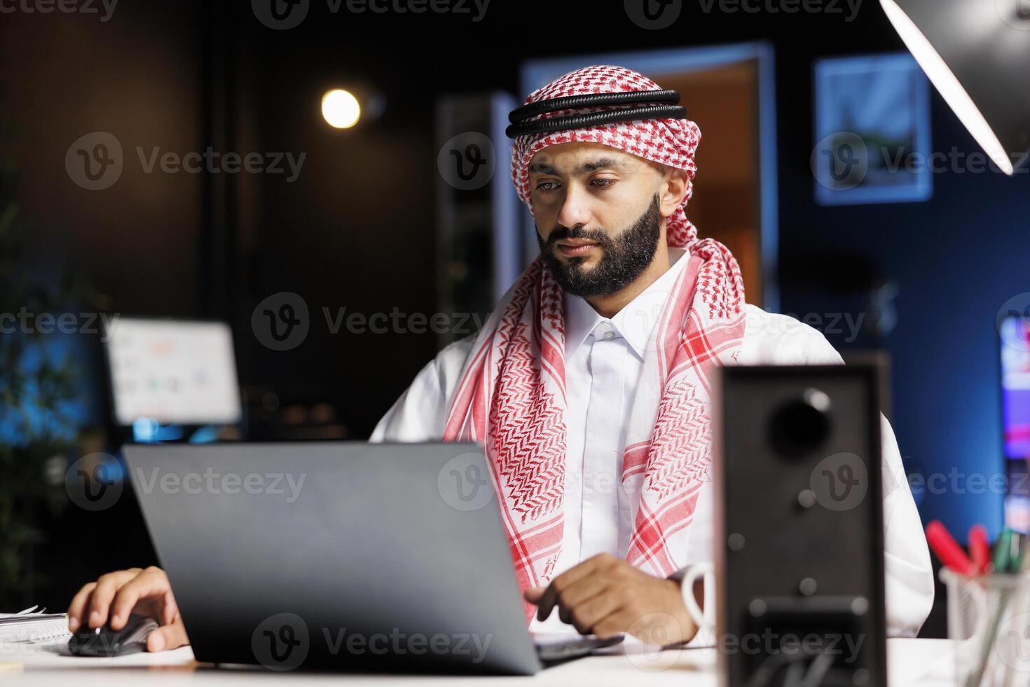 Arab businessman with confidence typing on a laptop at a contemporary desk. Male Muslim person using his personal computer for email and browsing in a serene and professional workplace environment. photo