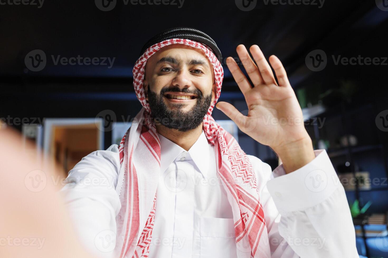 POV of a cheerful Muslim guy dressed in traditional clothing, engaging in call. Smiling Arab with thobe and headscarf grasping digital device and waving towards camera, doing online vlogging. photo