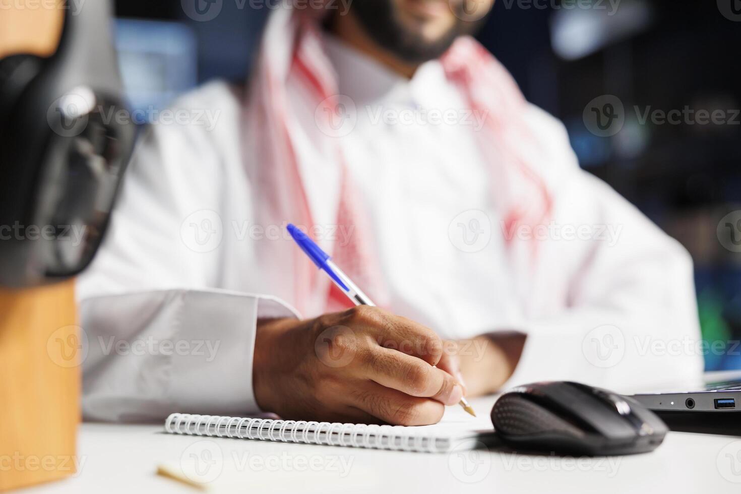 Close-up shot of a notebook and pen being used by a Middle Eastern man in traditional clothing seated at a table. Selective focus on Muslim guy writing down research notes. photo
