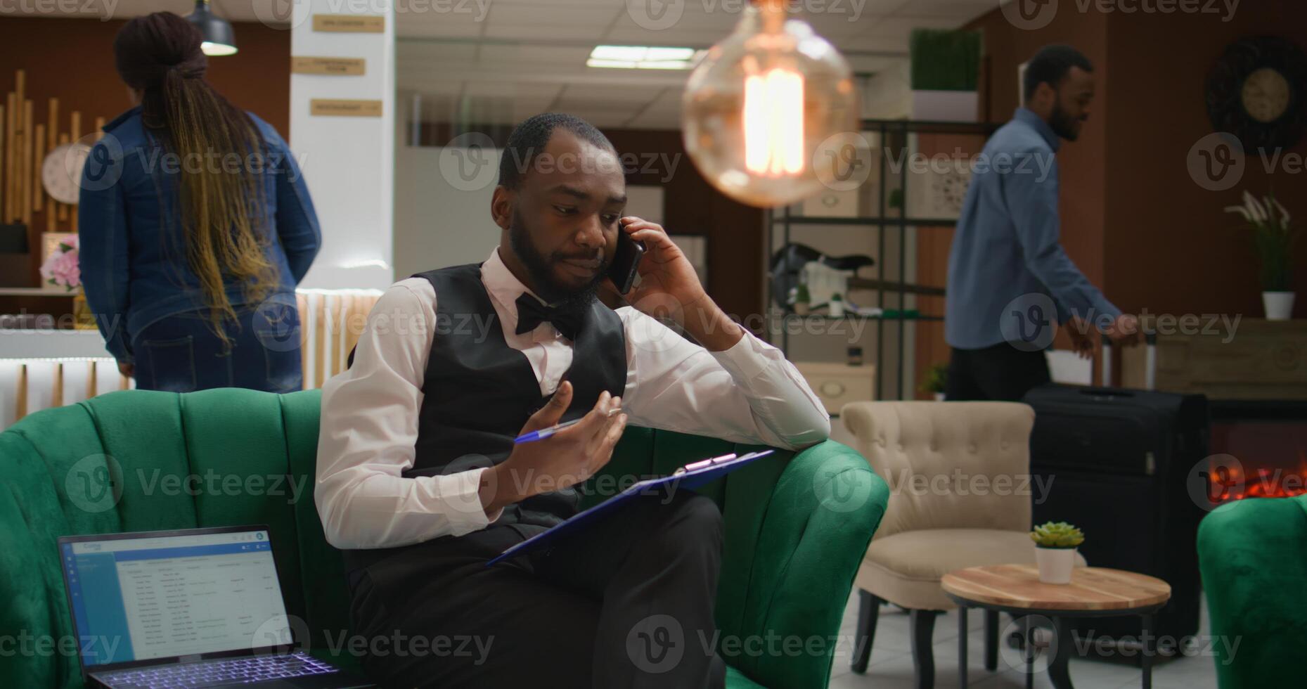 Bellhop talking on phone call and writing down information about reservation at hotel, working in tourism industry at five star resort. Employee calling guests to ensure safe check in. photo