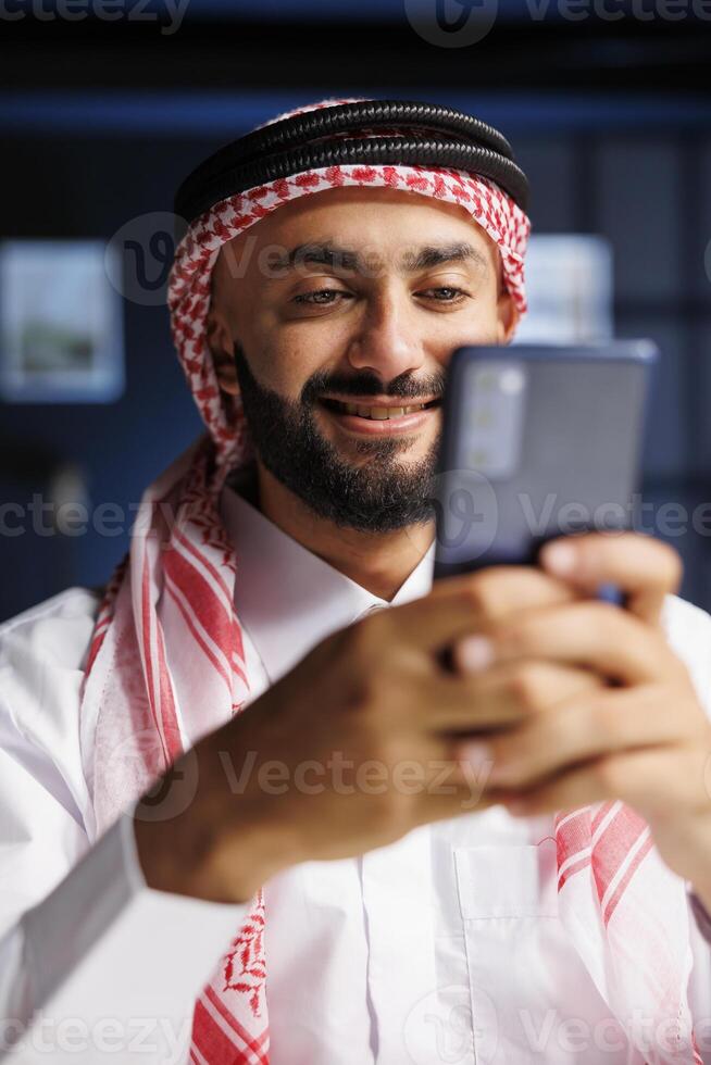 Detailed portrait shot of a smiling Muslim man using a smartphone for digital communication. Close-up view of an Arab entrepreneur typing and researching on his mobile device. photo