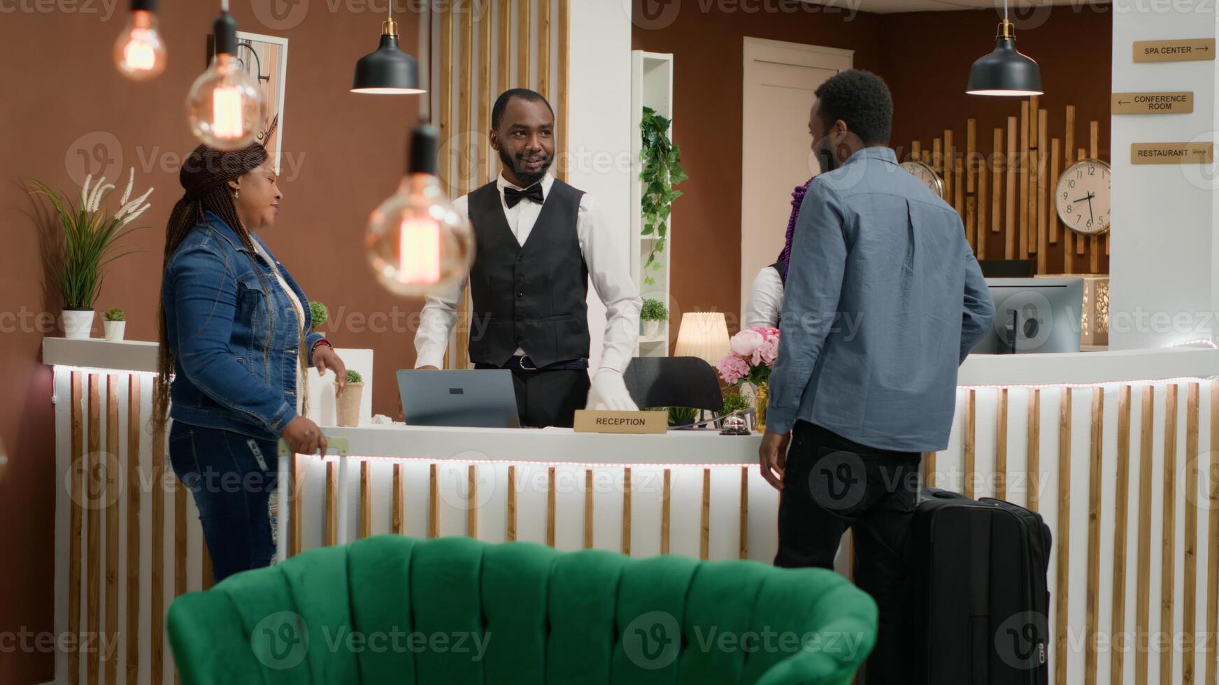 African american couple arrive at hotel, talking to front desk staff about room reservation and check in process. Man and woman entering lobby with trolley bags, travelling on honeymoon. photo