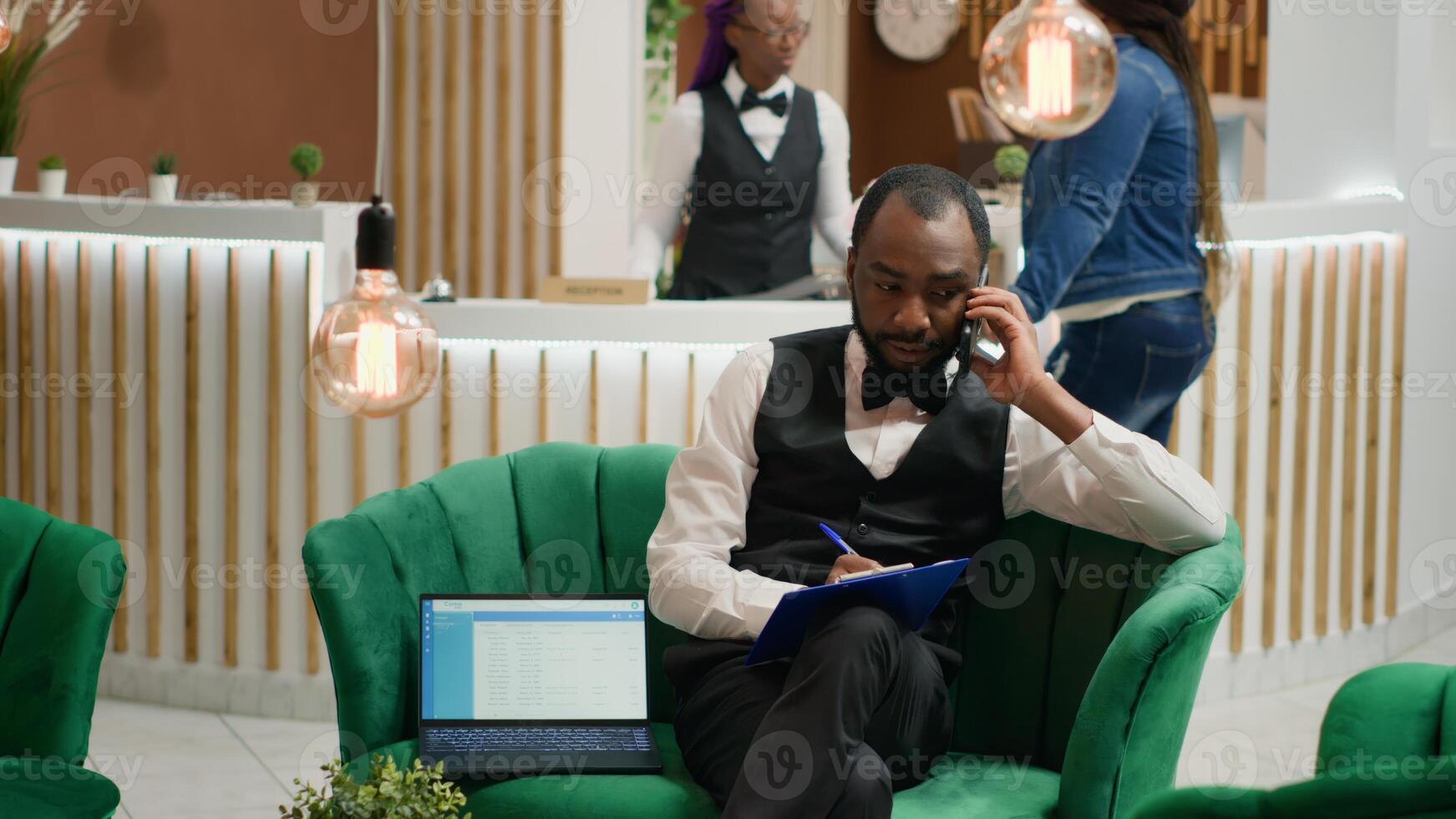 Bellboy talking on phone and taking notes, sitting in lobby at five star hotel. Concierge calling guests to ensure easy check in with all personal information, assisting visitors in arranging room. photo