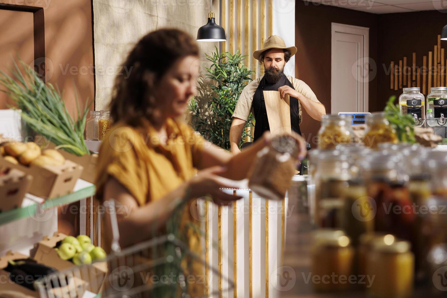 Zero waste shop storekeeper at checkout counter bagging organic bio vegetables grown locally, while customer in blurry foreground looks for pesticides free healthy groceries photo