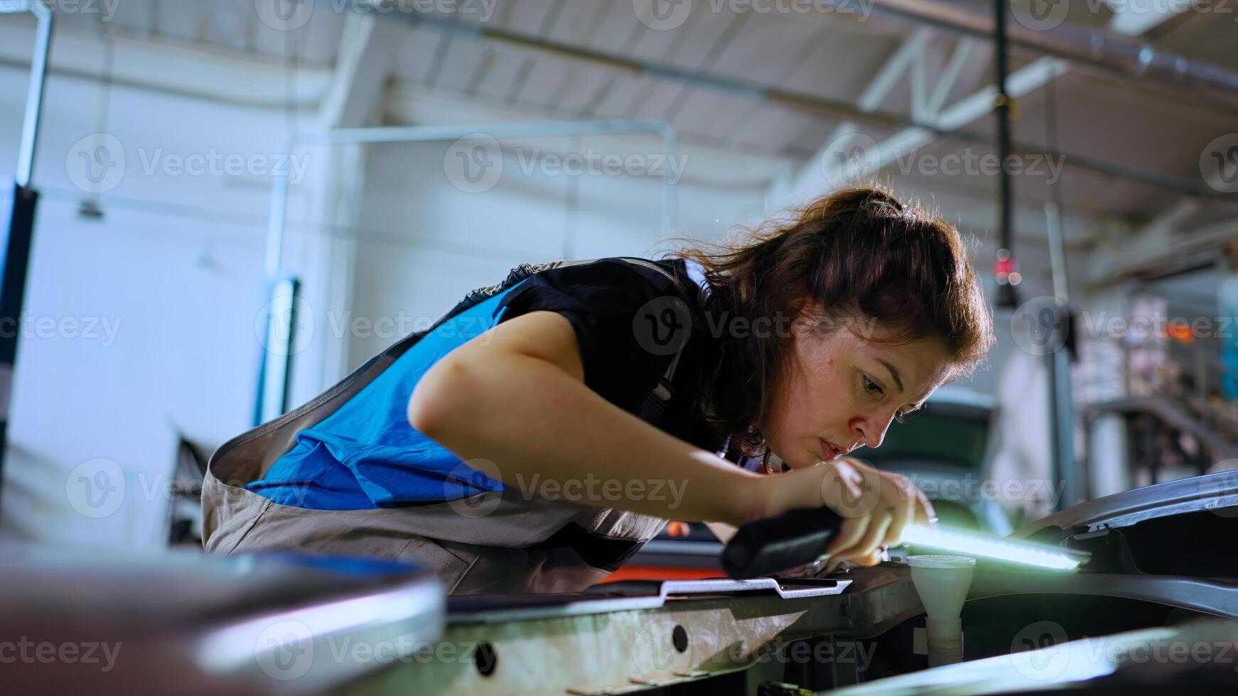 POV shot of mechanic opening car hood in auto repair shop, using work light to check for damages during routine maintenance. Garage workspace employee using professional tool to look inside vehicle photo