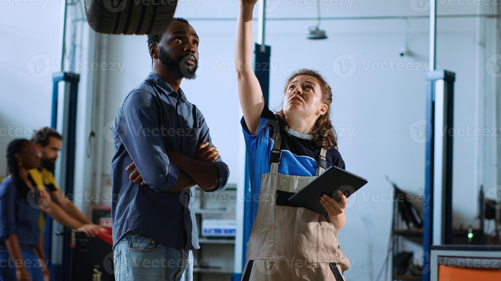 Mechanic in garage stands with customer underneath car on overhead lift, looking together for engine replacement. Expert helps BIPOC client recondition vehicle using tablet to find needed part photo