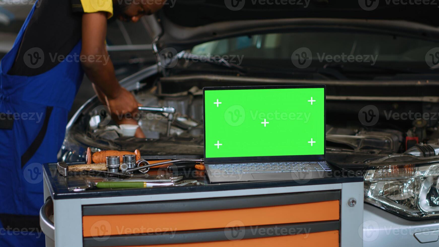 Close up shot of laptop placed on working bench in busy garage next to professional tools while serviceman fixes vehicle. Mockup device in auto repair shop with mechanic working in blurry background photo