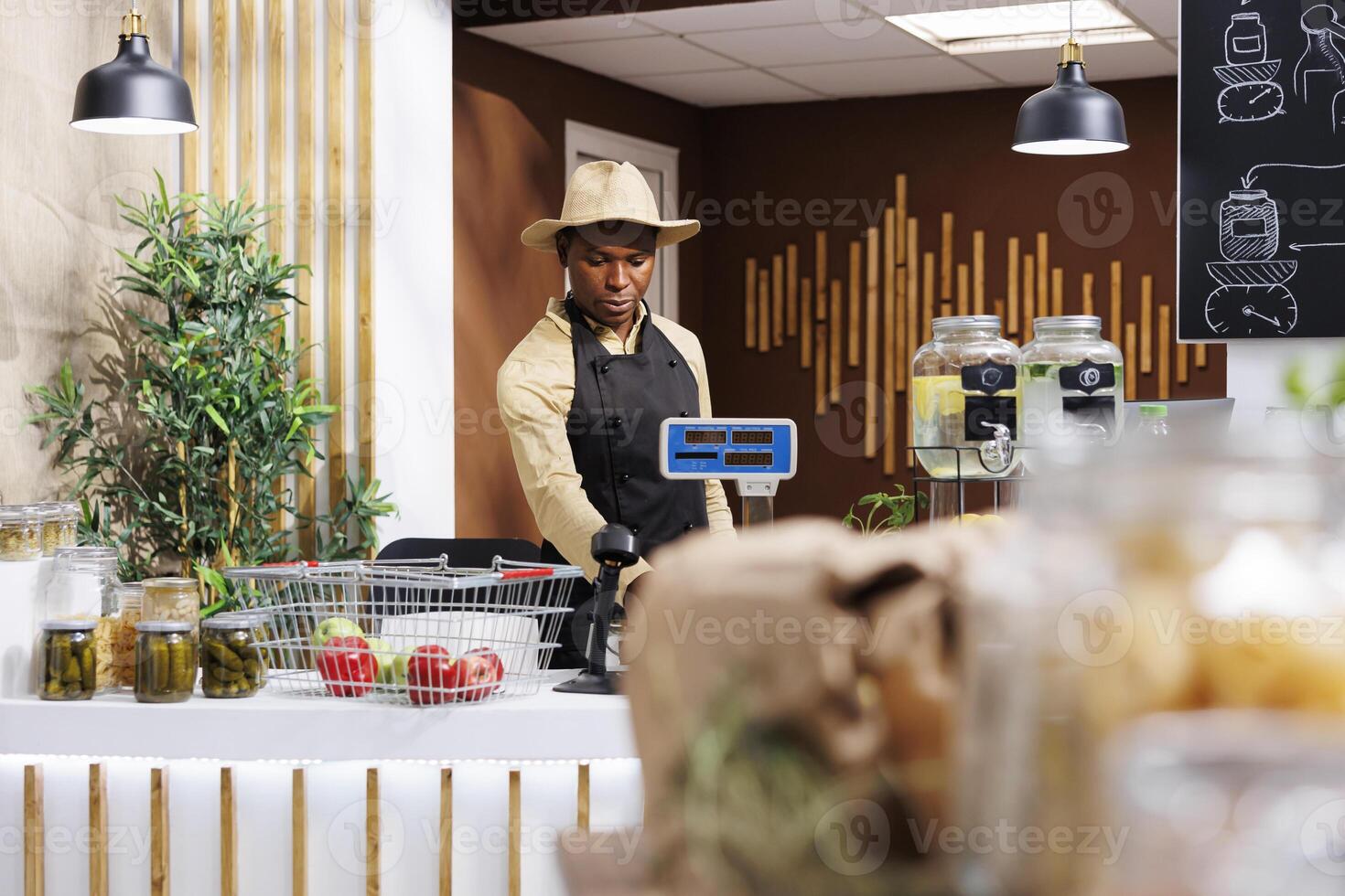 Focus on background of african american male shopkeeper at the checkout counter. Black man wearing a hat, arranging items over at the cashier desk in bio-store. Fresh, eco friendly produce displayed. photo